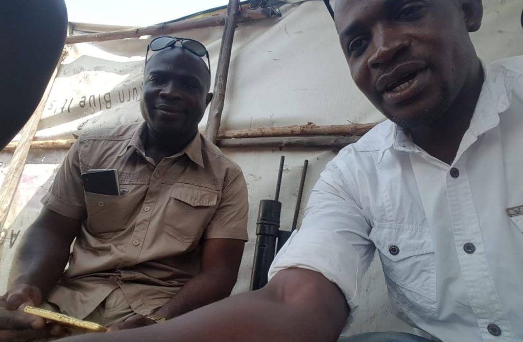 Inside a tent, two men are seated, with one man holding a gold block, symbolizing employees of the new Liberian mining company.